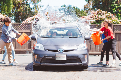 Three young people using buckets to throw water on a small sedan during a fundraising car wash