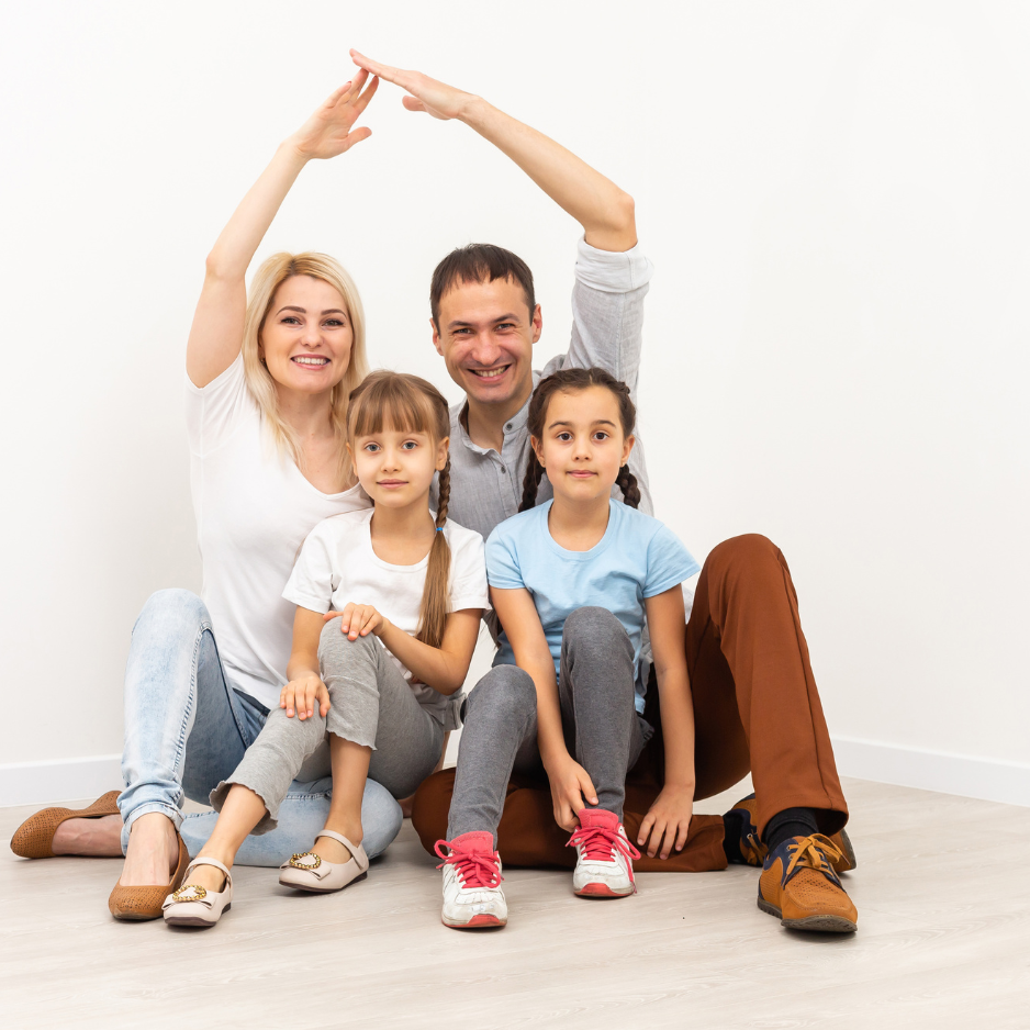 Young family, father, mother and two daughters sitting on the floor, while mother and father raise arms in the shape of a house roof